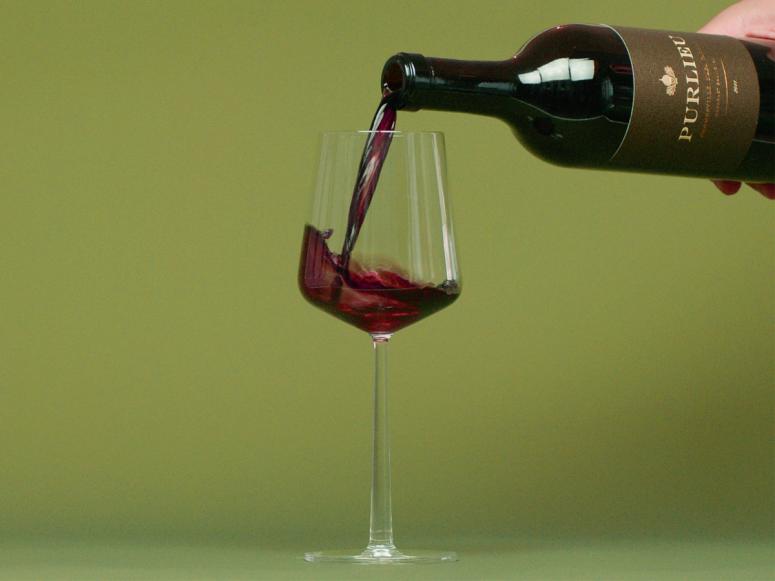 A hand pours a bottle of red wine into a wine glass on a green background.