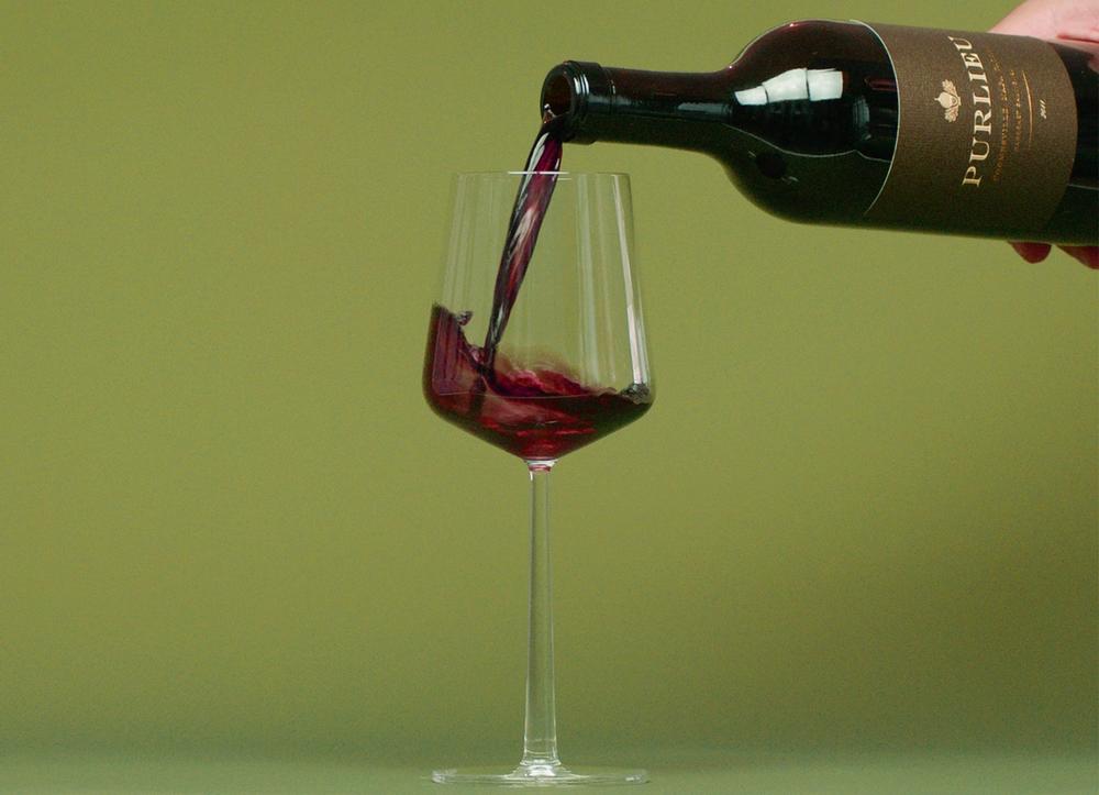 A hand pours a bottle of red wine into a wine glass on a green background.