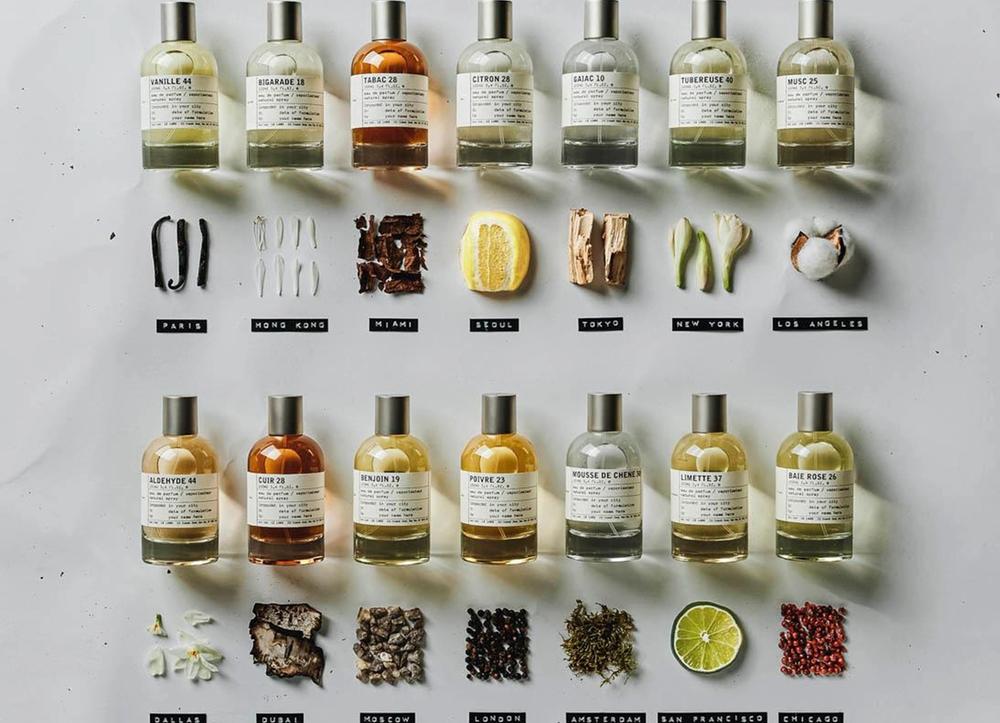 Fouteen Le Labo City Scents perfume bottles above an assortment of natural objects and their city names.