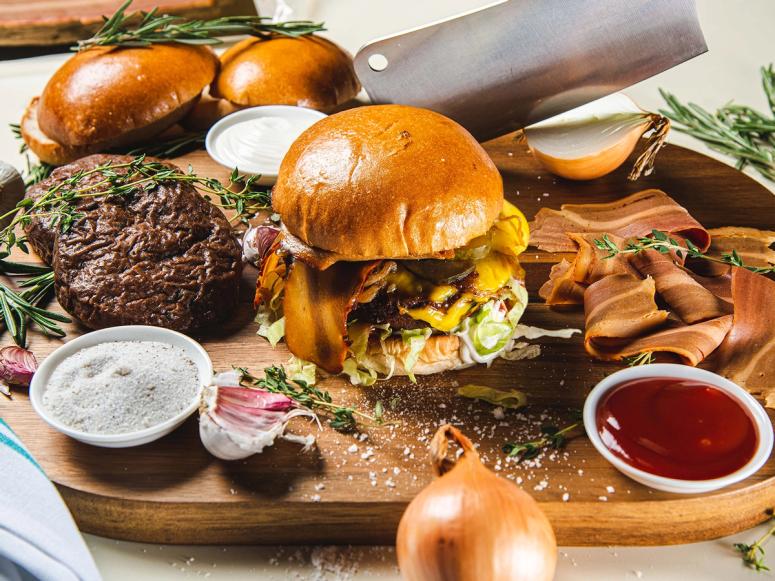 A carving board with vegan burgers and bacon, onions, a cleaver, herbs, and condiments.