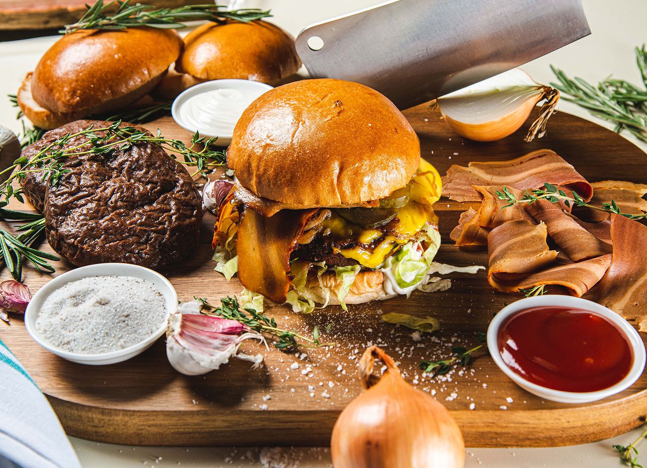 A carving board with vegan burgers and bacon, onions, a cleaver, herbs, and condiments.