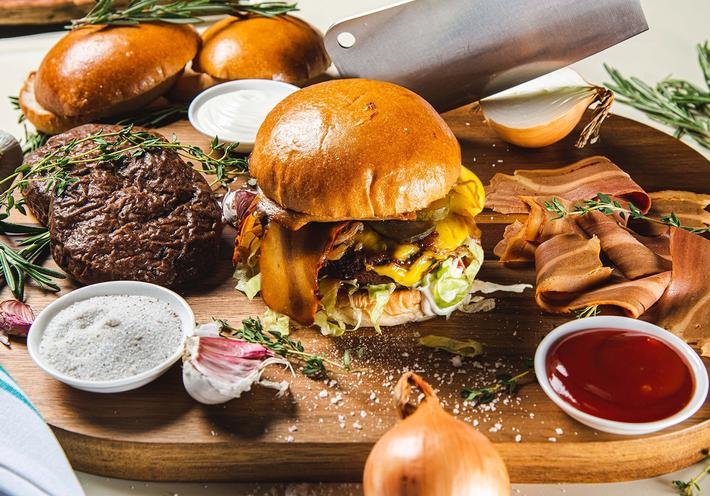A carving board with vegan burgers and bacon, onions, a cleaver, herbs, and condiments.
