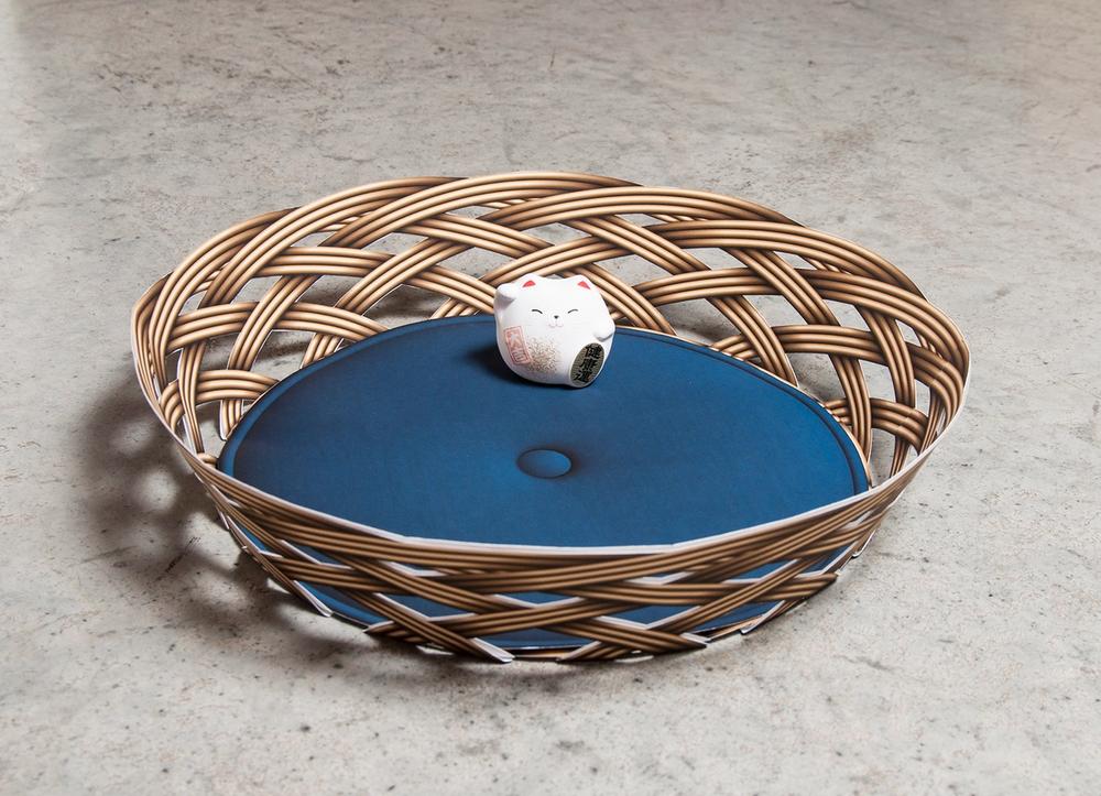 A round basket with a blue cushion and small cat figurine sitting inside.