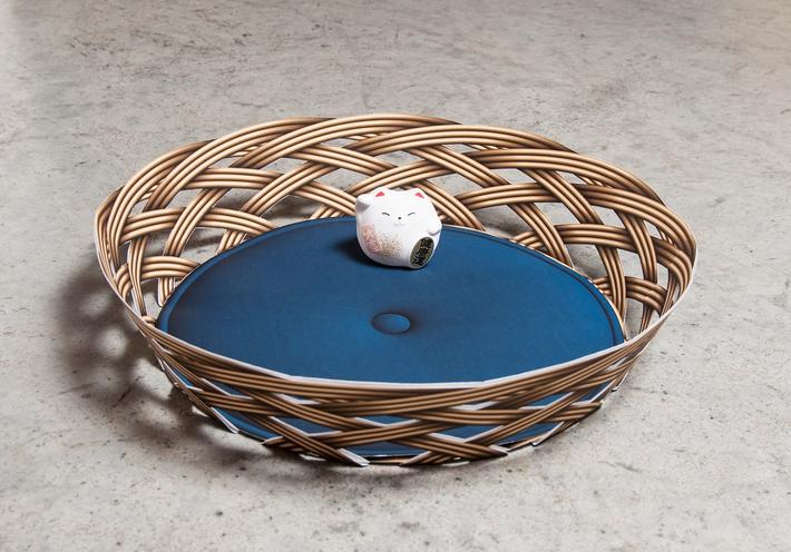 A round basket with a blue cushion and small cat figurine sitting inside.