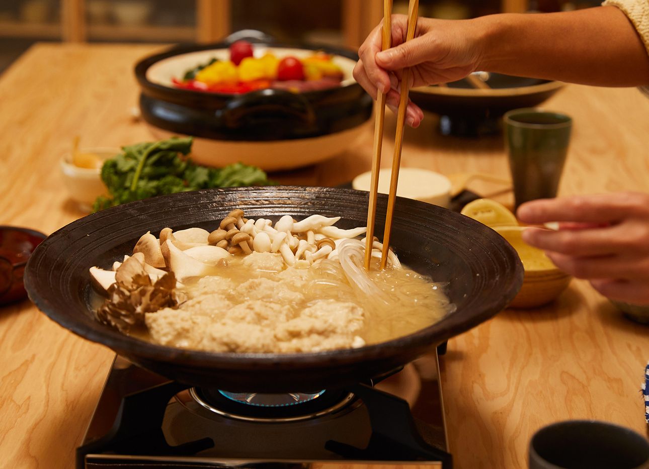 A donabe pot on a burner with mushrooms, noodles, and other ingredients in the background.