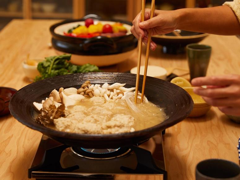 A donabe pot on a burner with mushrooms, noodles, and other ingredients in the background.