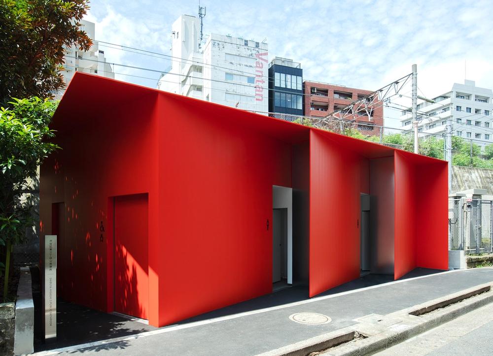 A bright red toilet building with angled entrances and the Tokyo cityscape in the background.