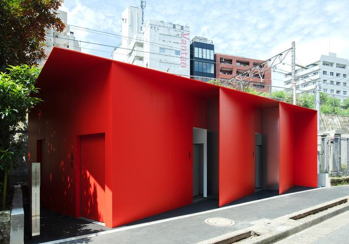 A bright red toilet building with angled entrances and the Tokyo cityscape in the background.