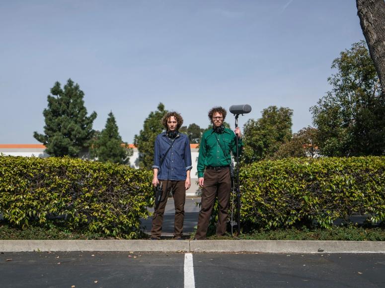 Sam Harnett and Chris Hoff holding audio equipment between two bushes in a parking lot.