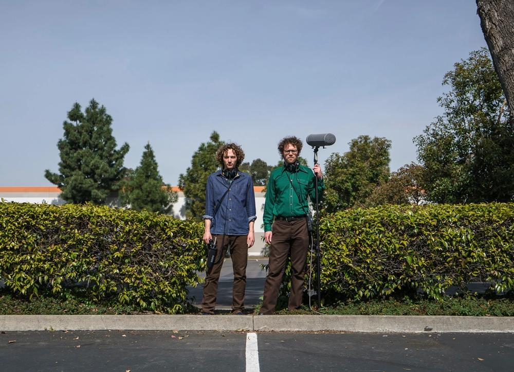 Sam Harnett and Chris Hoff holding audio equipment between two bushes in a parking lot.