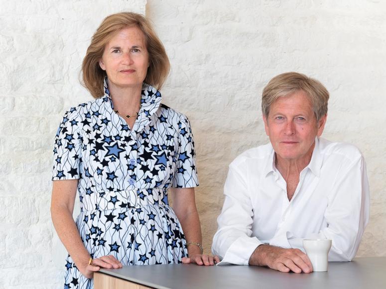 Catherine and John Pawson standing and siting at a table in front of a white wall.