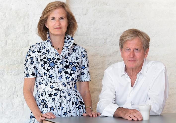 Catherine and John Pawson standing and siting at a table in front of a white wall.