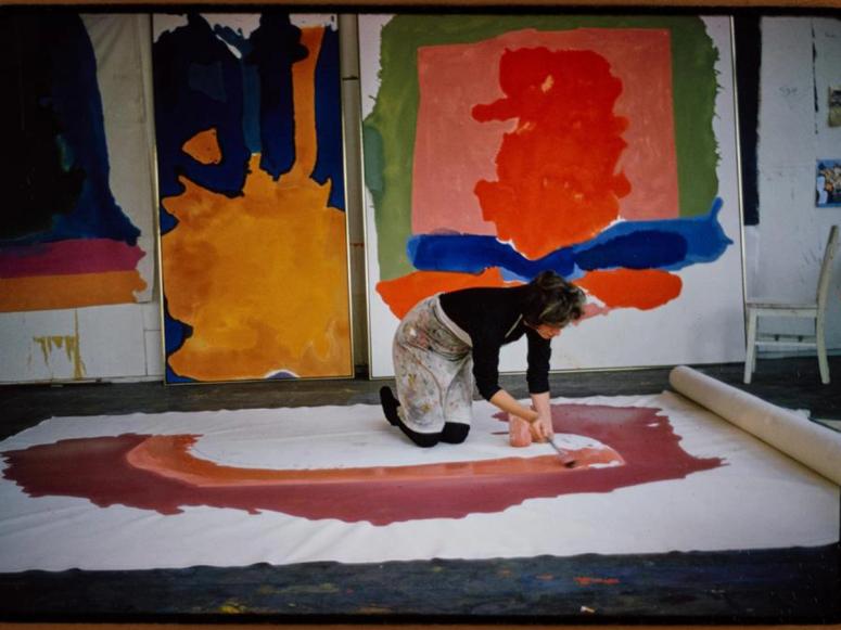 A woman kneels and paints a large rolled canvas on the ground, in front of other paintings lined up on a wall.