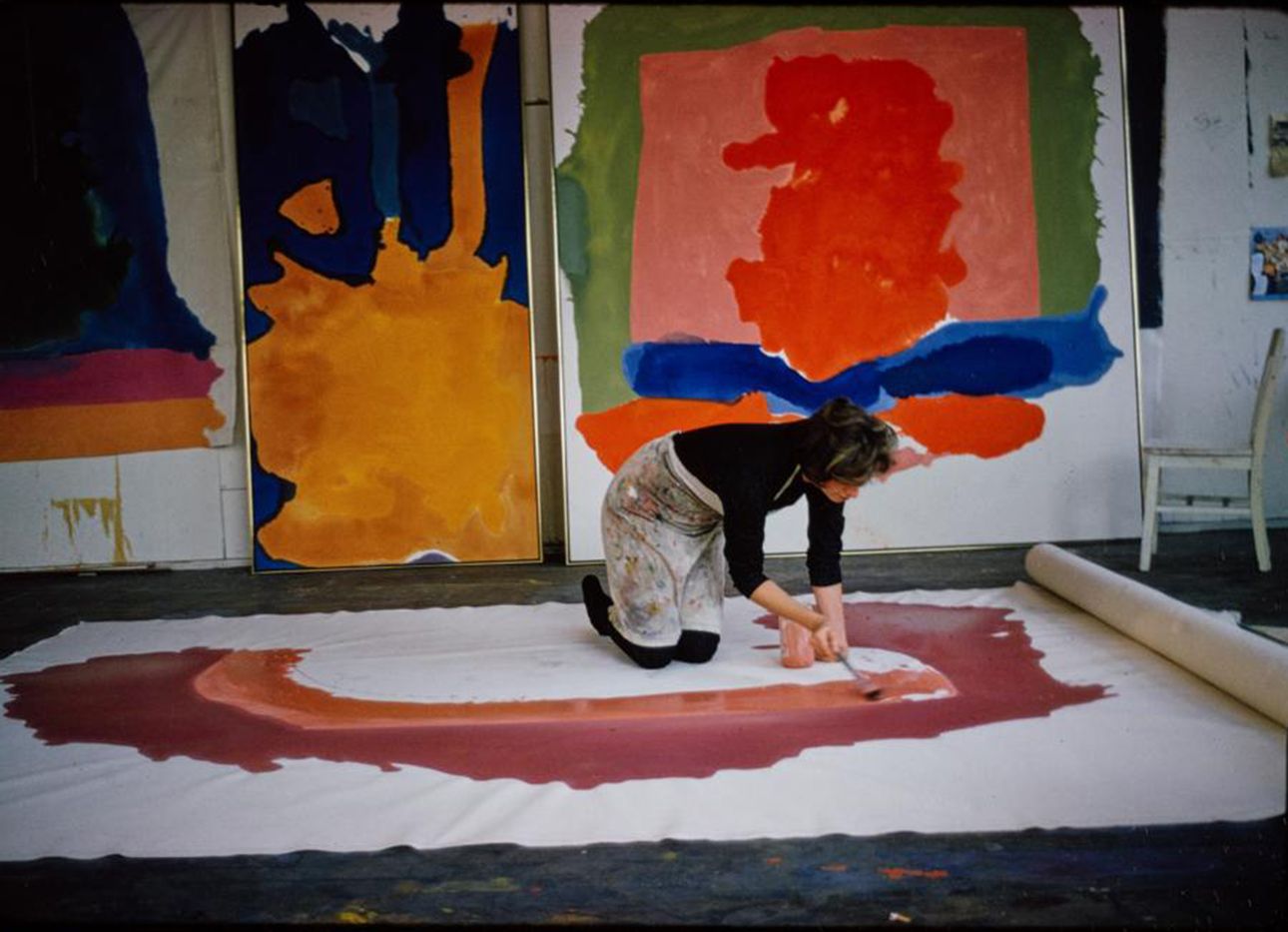 A woman kneels and paints a large rolled canvas on the ground, in front of other paintings lined up on a wall.