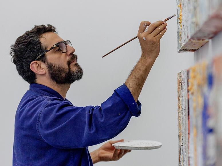 Jose Parla in a bright blue jacket and glasses, painting two canvases.