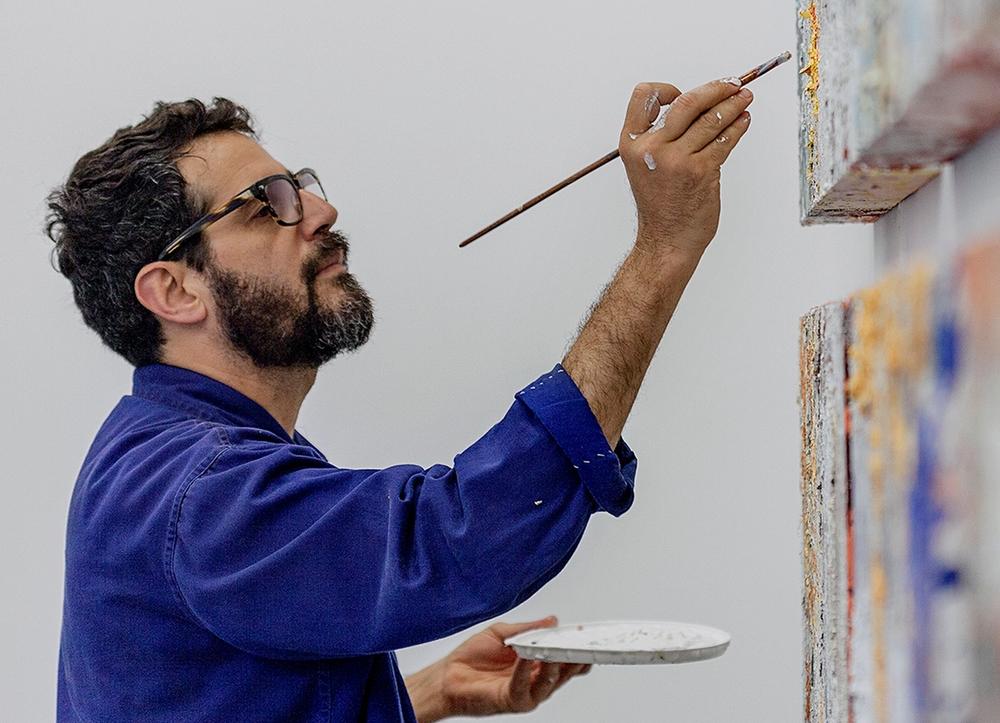 Jose Parla in a bright blue jacket and glasses, painting two canvases.