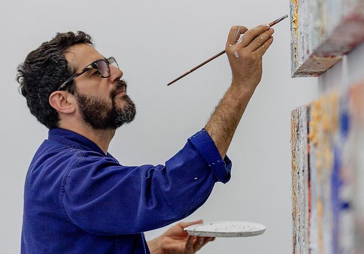 Jose Parla in a bright blue jacket and glasses, painting two canvases.