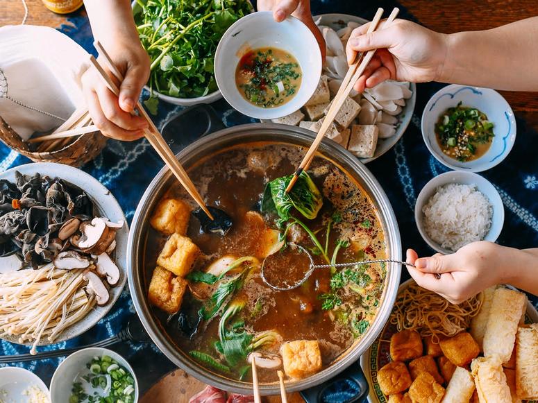 A large spread of dishes with three people adding various items to a hotpot.