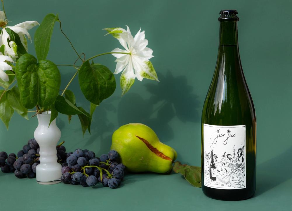 A bottle of Jus Jus next to a pear, purple grapes, and a white flower.