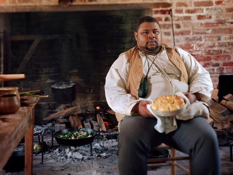 Michael W. Twitty sitting with a plate of biscuits in front of a large open fireplace.