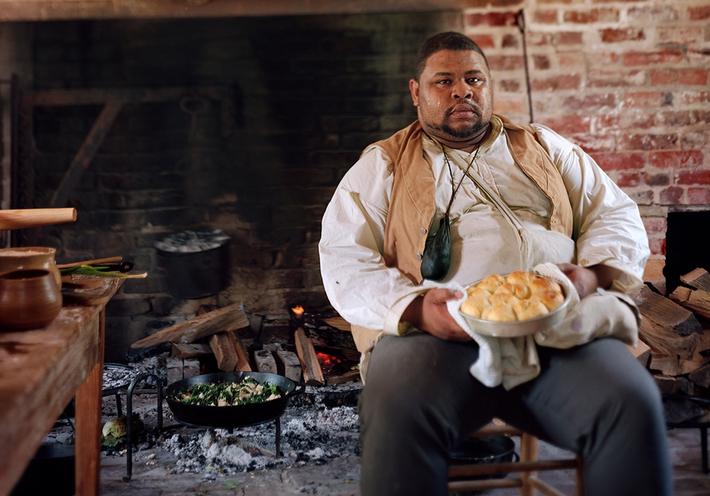 Michael W. Twitty sitting with a plate of biscuits in front of a large open fireplace.