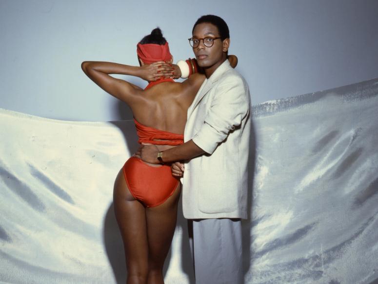 Willi Smith looking into the camera, his hand on a model with a red bathing suit, facing away.