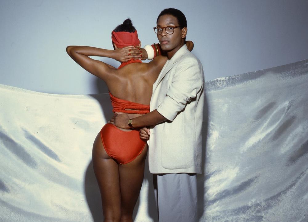 Willi Smith looking into the camera, his hand on a model with a red bathing suit, facing away.