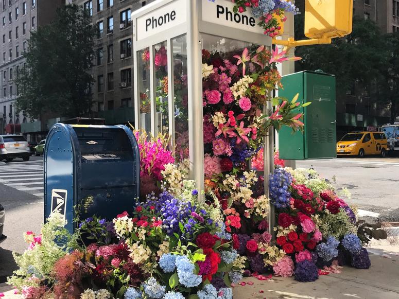 Hundreds of flowers spill out of a phone booth on on New York City street corner.