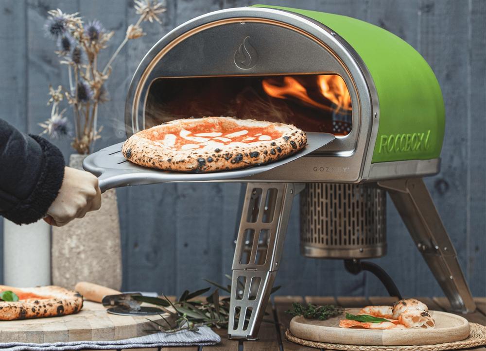 A hand pulling a steaming margherita pizza out of a green Roccbox oven.