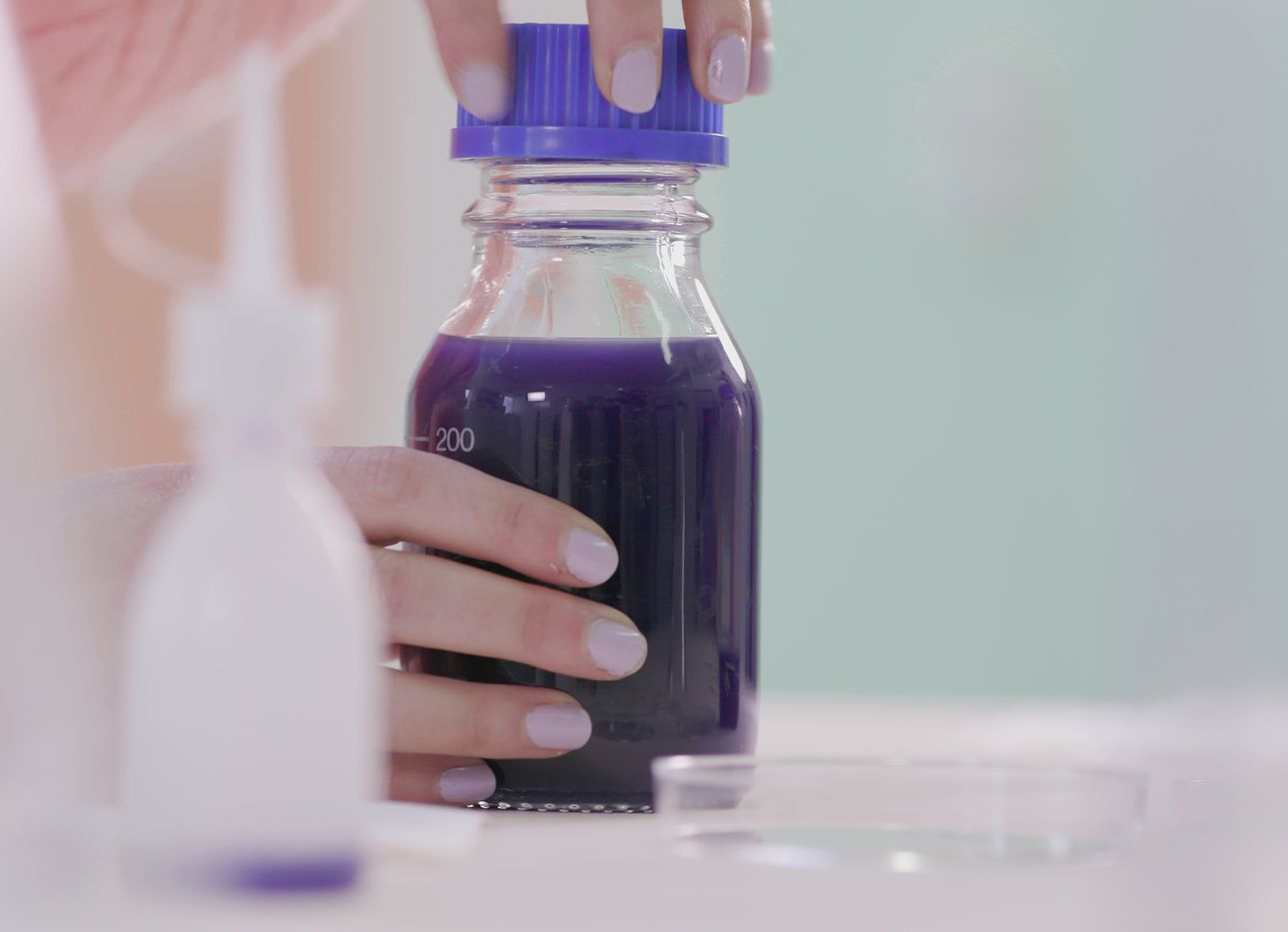 Two hands with purple nails holding a beaker filled with purple liquid.