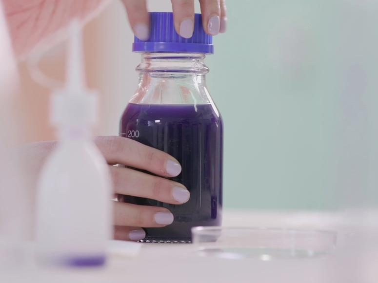 Two hands with purple nails holding a beaker filled with purple liquid.