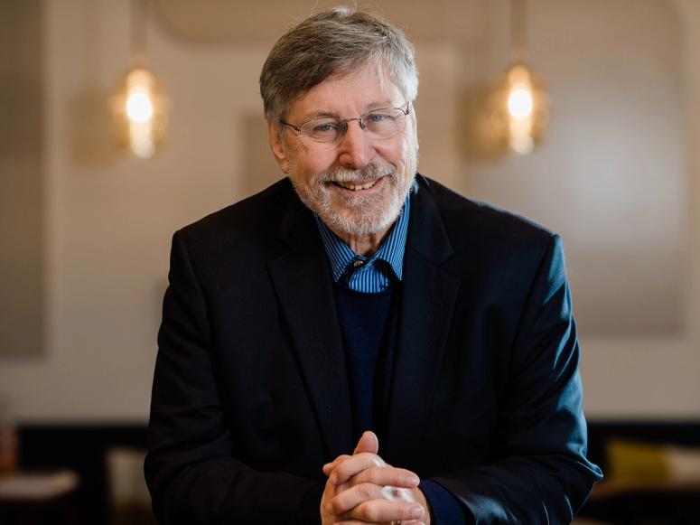 Dr. Bessel van der Kolk in a dark suit with his hands clasped, smiling.