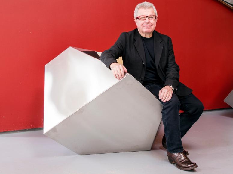 Daniel Libeskind in a black suit in front of a red wall, sitting on a geometric seat.