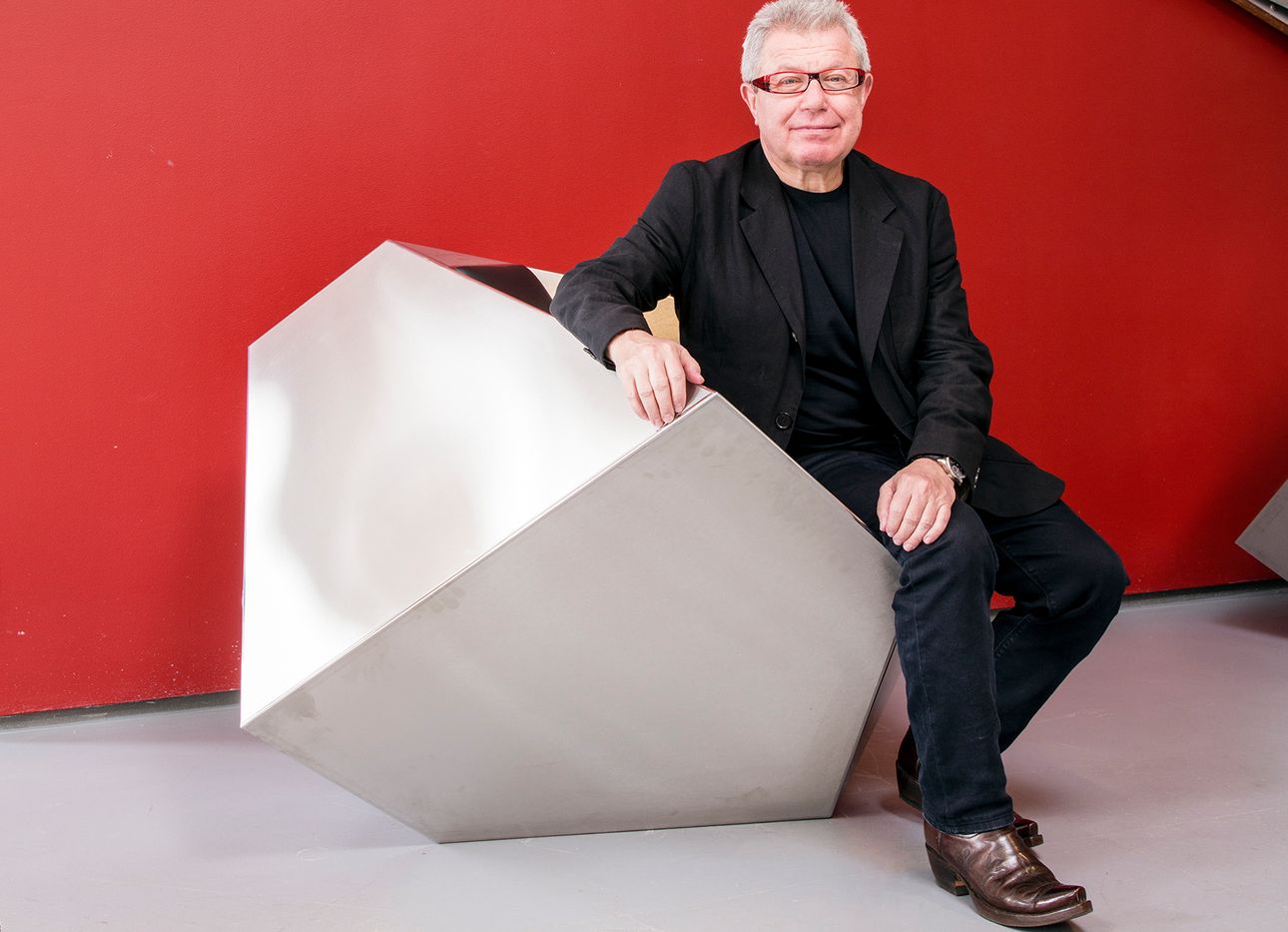 Daniel Libeskind in a black suit in front of a red wall, sitting on a geometric seat.