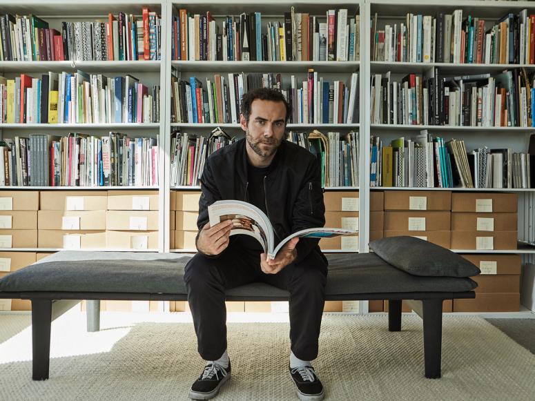 Jonathan Olivares sitting on a daybed in his library, reading a large art book.