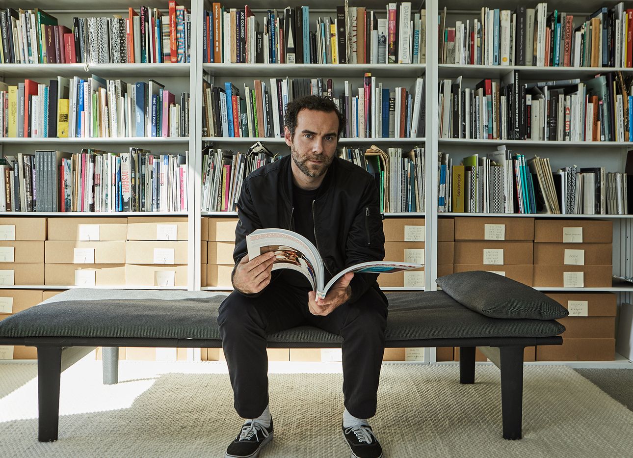 Jonathan Olivares sitting on a daybed in his library, reading a large art book.