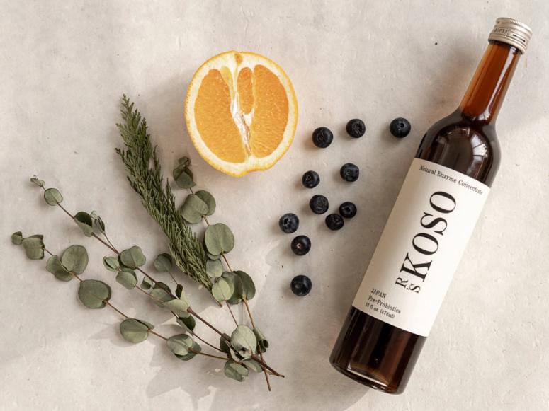 A bottle of R's Koso next to blueberries, half an orange, and herbs.