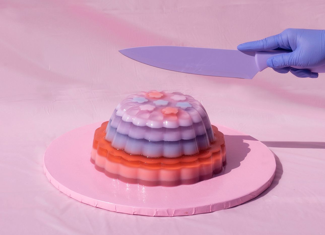 A hand in a blue plastic glove holds a purple knife above a pink, purple, and red jelly cake atop a pink platter.