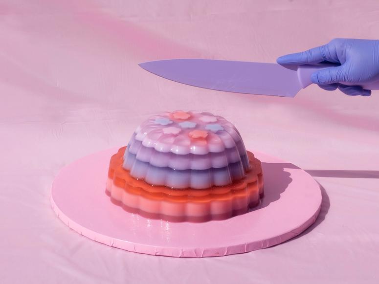 A hand in a blue plastic glove holds a purple knife above a pink, purple, and red jelly cake atop a pink platter.