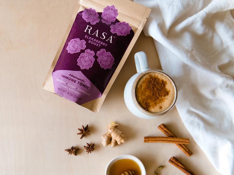 A cup of Rasa Elderberry Boost with cloves of cinammon, ginger, and anise on a bright wood table.