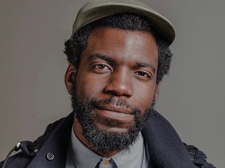 Steven Satterfield in a hat and jacket, on a gray background.
