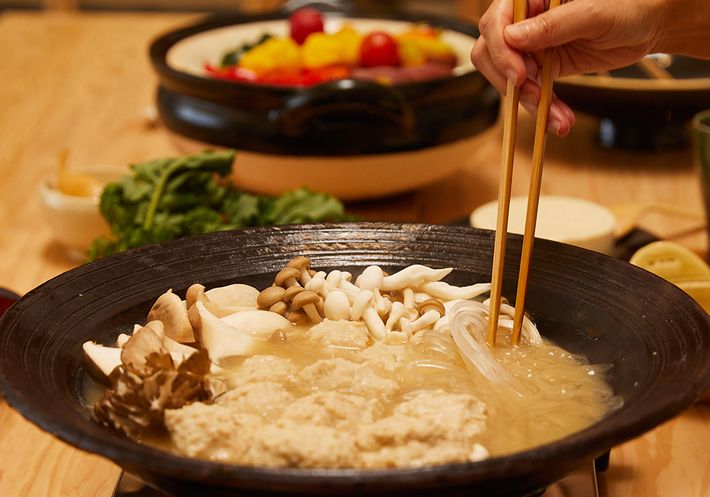 A donabe pot on a burner with mushrooms, noodles, and other ingredients in the background.