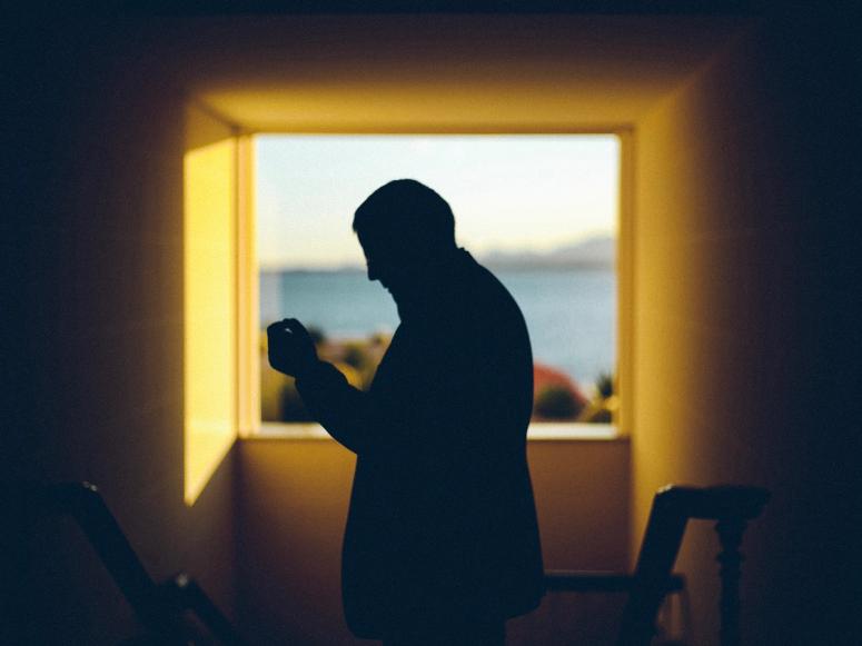 A man's silhouette sniffs perfume near a bright window.