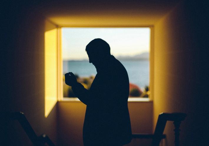 A man's silhouette sniffs perfume near a bright window.