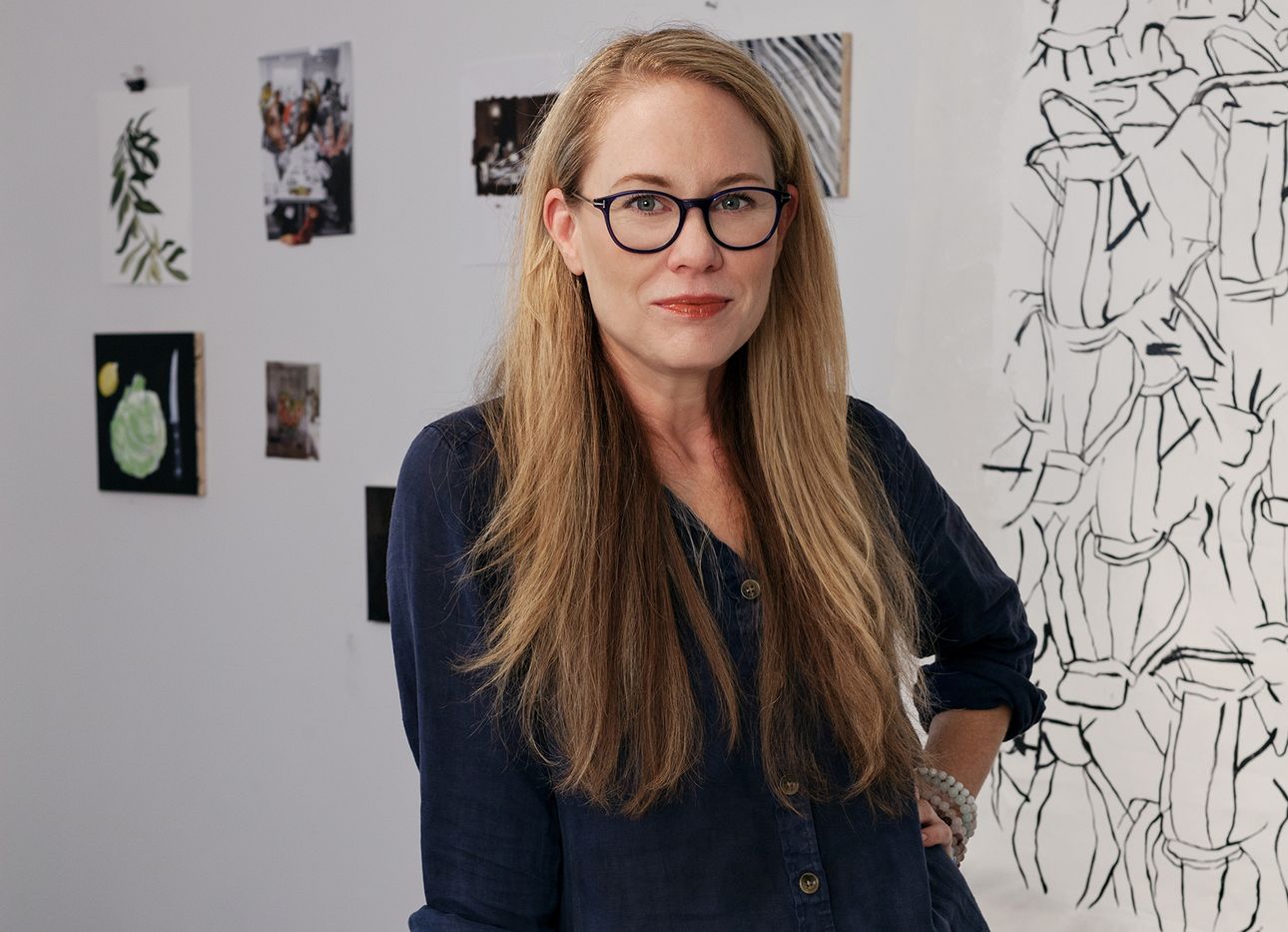 Catherine Haley Epstein stands with her hand on her hip in front of several drawings.