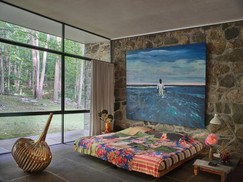 A bedroom at the Eliot Noyes House, with a large blue painting, multicolored bedspread, and brass sculpture near a large glass window.