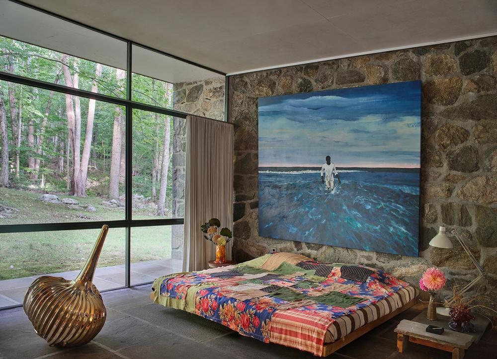 A bedroom at the Eliot Noyes House, with a large blue painting, multicolored bedspread, and brass sculpture near a large glass window.