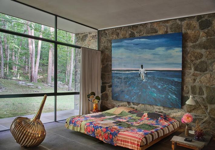 A bedroom at the Eliot Noyes House, with a large blue painting, multicolored bedspread, and brass sculpture near a large glass window.