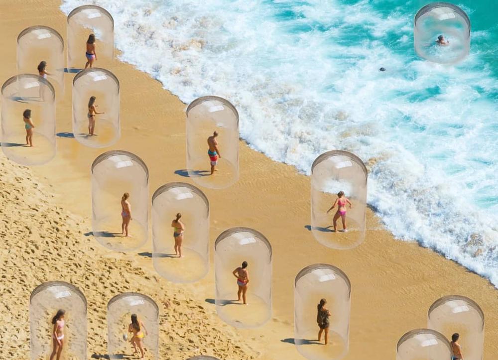 Various people encased in glass bubbles stand on the beach near the ocean