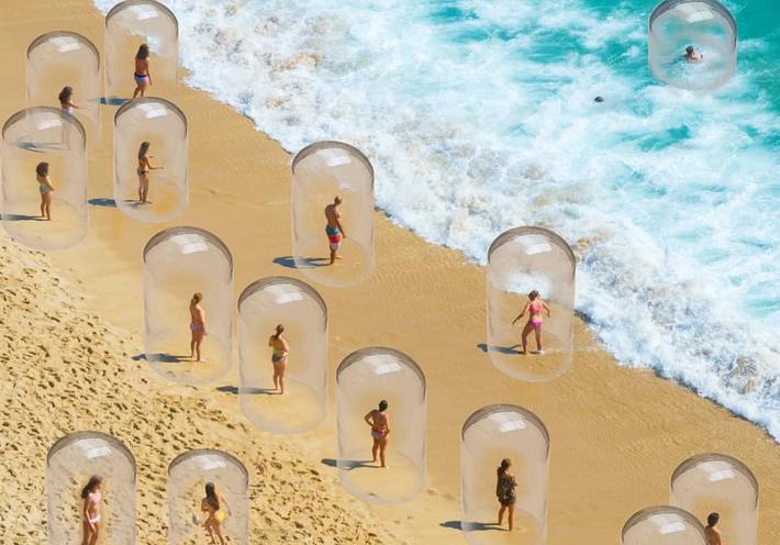 Various people encased in glass bubbles stand on the beach near the ocean